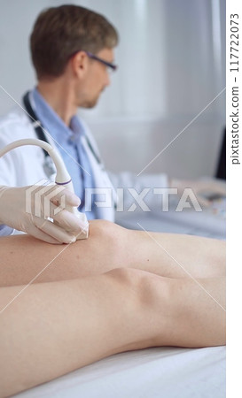 Doctor man with blue protective gloves using ultrasound equipment on the knee of a female patient, analyzing the results on a monitor in the background. Medicine 117722073