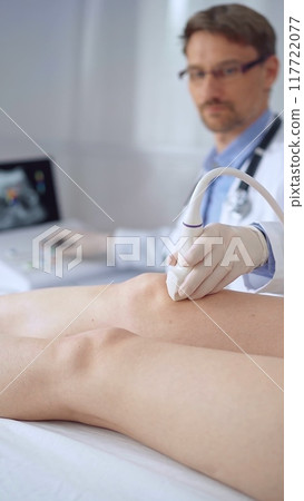 Doctor man with blue protective gloves using ultrasound equipment on the knee of a female patient, analyzing the results on a monitor in the background. Medicine 117722077