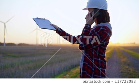 Female engineer talking on the phone in a field with wind turbines, as the sun sets. Concept of clean energy 117722088