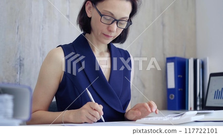 Businesswoman doing paperwork at her desk in the office, using calculator and taking notes. Taxes, audit in business Businesswoman doing paperwork at her desk in the office, using calculator and taking notes. Taxes, audit in business 117722101