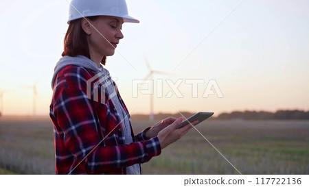 Woman engineer taking notes with a tablet computer on a field with wind turbines, as the sun sets. Clean energy and engineering concept 117722136