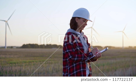 Adult woman engineer taking notes on a clipboard on a field with wind turbines, as the sun sets Adult woman engineer taking notes on a clipboard on a field with wind turbines, as the sun sets 117722137