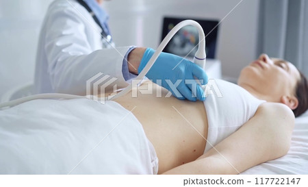 Doctor man, wearing blue medical gloves, is using ultrasound equipment on the breast of a female patient lying down for a medical cardiology examination. Medicine concept Doctor man, wearing blue medical gloves, is using ultrasound equipment on the breast of a female patient lying down for a medical cardiology examination. Medicine concept 117722147