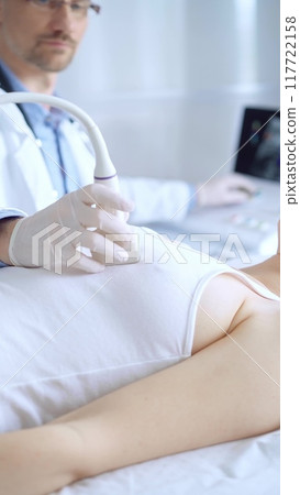 Doctor man wearing white protective gloves is using ultrasound equipment on the breast of a female patient. Mammary gland health. Medicine concept 117722158