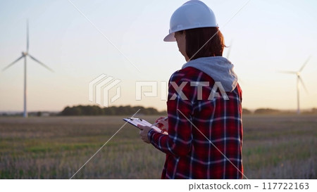 Adult woman engineer wearing white cask is taking notes on a clipboard on a field with wind turbines Adult woman engineer wearing white cask is taking notes on a clipboard on a field with wind turbines 117722163