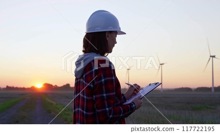 Woman engineer is taking notes on a clipboard on a field with wind turbines, as the sun sets in evening. Clean energy and engineering 117722195