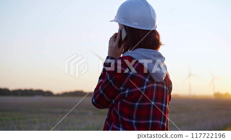 Woman engineer talking on the phone in a field with wind turbines, as the sun sets. Concept of clean energy and engineering Woman engineer talking on the phone in a field with wind turbines, as the sun sets. Concept of clean energy and engineering 117722196