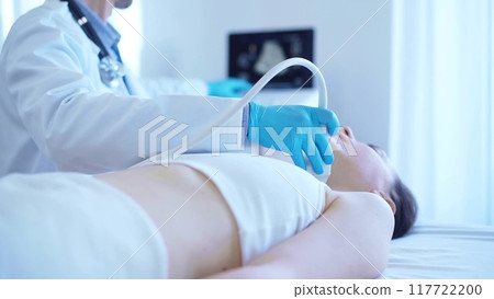 Doctor man with blue medical gloves is using ultrasound machine to examine neck of female patient lying down in clinic procedure cabinet. Medicine service 117722200