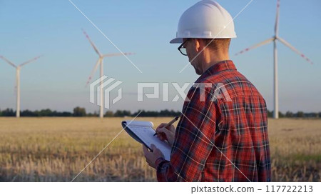 Man engineer, wearing a white protective helmet is taking notes with a clipboard in a field with wind turbines, as the sun sets. Clean energy and engineering concept Man engineer, wearing a white protective helmet is taking notes with a clipboard in a field with wind turbines, as the sun sets. Clean energy and engineering concept 117722213