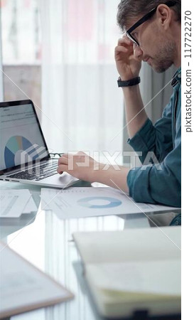 Businessman wearing green shirt and glasses is working with laptop computer and data analysis charts in office. Vertical business portrait 117722270