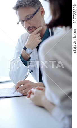 Doctor and a patient. The physician, wearing a white medical coat over a green shirt, gesturing with his hands during a consultation in the clinic. Medicine concept 117722296