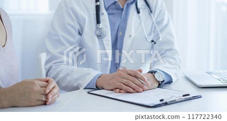 A doctor and a patient. The physician, wearing a white medical coat over a blue shirt, is filling out a medical record form during a consultation in the clinic. Medical service 117722340