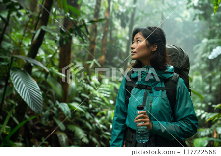 Young woman traveler dressed in a green rain jacket hiking through a dense alone with backpack and bottle of water. Local single travel practices 117722568
