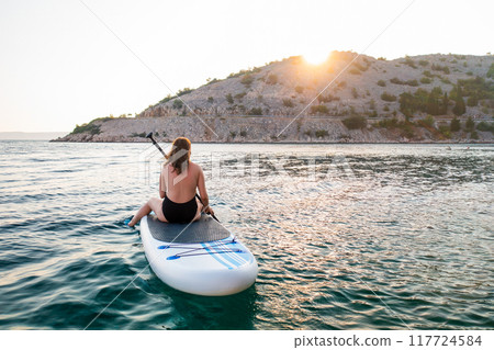 A woman enjoys paddle boarding on calm waters at sunset near a scenic coastal landscape 117724584