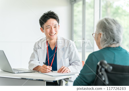 A young Asian male doctor (family doctor) smilingly examining and questioning an elderly woman at a hospital or nursing home 117726283