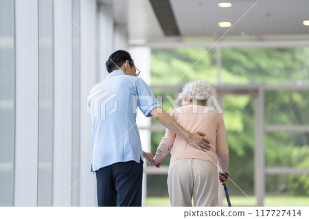 Rear view of a senior woman walking with a cane and a caregiver 117727414