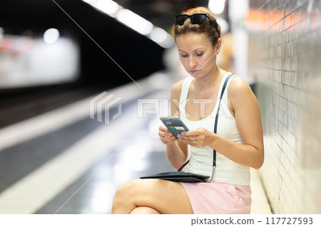 Adult woman sits at subway station with phone 117727593