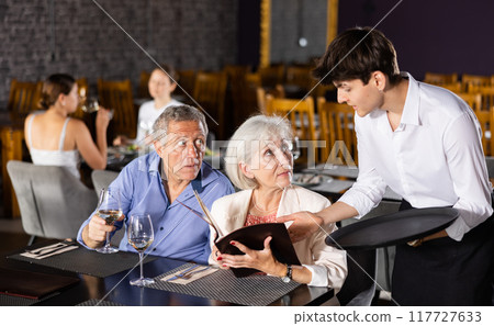 Young male waiter takes order from elderly couple in restaurant Young male waiter takes order from elderly couple in restaurant 117727633