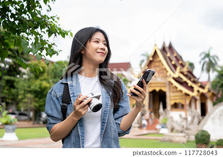An attractive Asian female traveler holding a camera and smartphone, visits a Thai temple. An attractive Asian female traveler holding a camera and smartphone, visits a Thai temple. 117728043