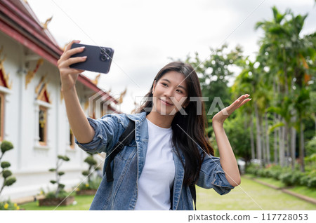 A joyful Asian female tourist is taking a picture or video of herself in a stunning Thai temple. 117728053