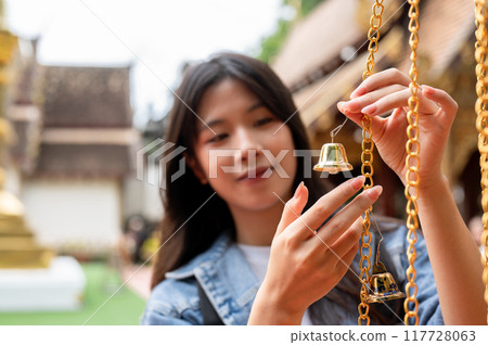 A positive Asian female tourist is hanging a bell for luck in a temple during her visit to Thailand. 117728063