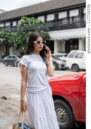 A fashionable Asian woman is talking on the phone while walking by the street in the city. A fashionable Asian woman is talking on the phone while walking by the street in the city. 117728106