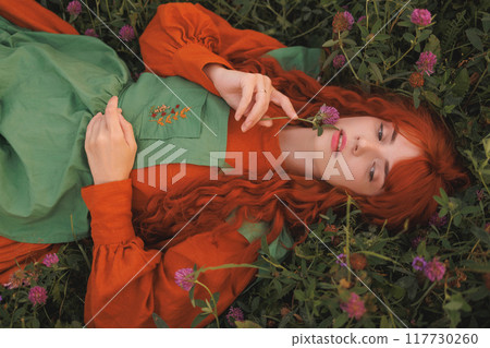 A shepherdess woman with red hair and a green dress sits in a field of flowers. A shepherdess woman with red hair and a green dress sits in a field of flowers. 117730260