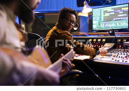 Musician composing a new record with his guitar and mixing console in studio, collaborating with music producer to create sounds. Audio technician uses control panel board to edit tracks. Musician composing a new record with his guitar and mixing console in studio, collaborating with music producer to create sounds. Audio technician uses control panel board to edit tracks. 117730901
