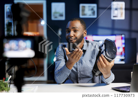 Smiling tech content creator filming technology review of newly released VR glasses, unboxing them and presenting specifications to audience. Influencer showing virtual reality device to subscribers Smiling tech content creator filming technology review of newly released VR glasses, unboxing them and presenting specifications to audience. Influencer showing virtual reality device to subscribers 117730973