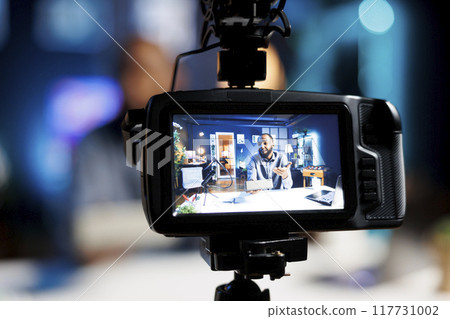 Focus on professional video production camera used for capturing footage of audio engineer in blurry background filming tech reviews for online audience, showcasing Bluetooth portable speaker 117731002