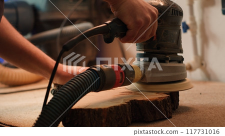 Craftsperson works with orbital sander on lumber block to achieve smooth finish, close up shot. Woodworking specialist in furniture assembly shop using angle grinder to fix wooden surface, camera A Craftsperson works with orbital sander on lumber block to achieve smooth finish, close up shot. Woodworking specialist in furniture assembly shop using angle grinder to fix wooden surface, camera A 117731016