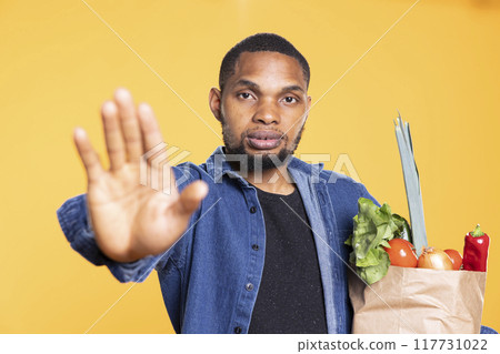 Stressed serious man raising his palm and doing stop symbol, asking for a break against yellow background. Young male model carrying a groceries paper bag, prohibiting something. 117731022