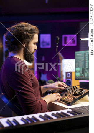 Young musician composing soundtracks and editing them on mixing console, creating cool tunes on daw software interface in his home studio. Producer adjusting volume levels on stereo gear. 117731027