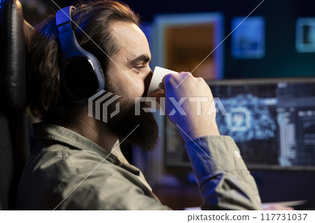 IT specialist drinking coffee with headphones on listening to songs, scripting artificial intelligence on computer. Developer enjoying hot beverage and music in ears while working on coding AI on PC IT specialist drinking coffee with headphones on listening to songs, scripting artificial intelligence on computer. Developer enjoying hot beverage and music in ears while working on coding AI on PC 117731037