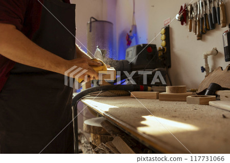 Manufacturer at workbench using manual sandpaper to sander lumber block, assembling furniture in studio. Carpenter smoothing piece of wood, sawdust flying in sunlight, close up shot Manufacturer at workbench using manual sandpaper to sander lumber block, assembling furniture in studio. Carpenter smoothing piece of wood, sawdust flying in sunlight, close up shot 117731066