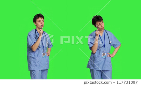 Medical assistant thinking about new treatment ideas in studio, acting thoughtful while she stands against greenscreen backdrop. Pensive young nurse contemplating about healthcare ideas. Camera B. 117731097