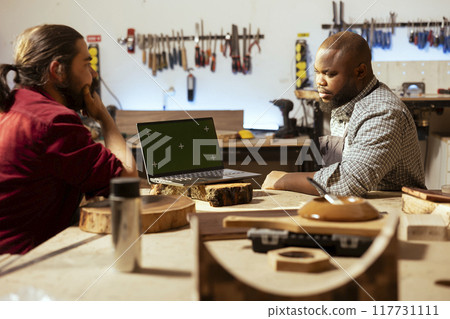 Manufacturers using CAD software on green screen laptop, debating wood design to use. Carpenters in assembly shop chatting, using program on mockup notebook to make furniture schematics 117731111