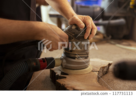 Artisan in studio enjoying diy hobby, using orbital sander with fine sandpaper on lumber to achieve smooth finish. Creative person in carpentry shop uses angle grinder to create wooden objects Artisan in studio enjoying diy hobby, using orbital sander with fine sandpaper on lumber to achieve smooth finish. Creative person in carpentry shop uses angle grinder to create wooden objects 117731113