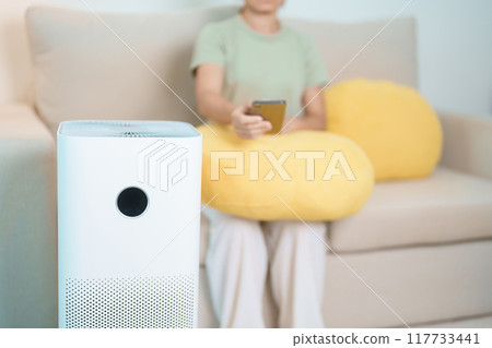 Woman turn on Air Purifier in living room by smartphone application. Purification system for filter and cleaning dust PM2.5 HEPA and virus. Allergy, Pure air, health lifestyle and Air Pollution 117733441