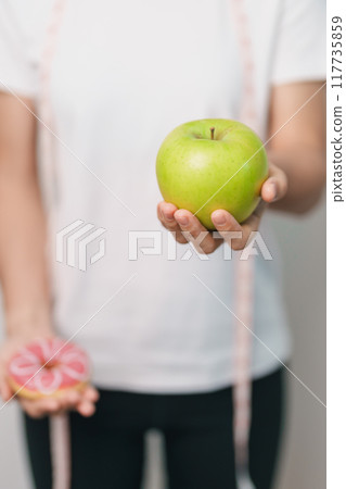 woman hand hold green Apple and donut, female fitness choose between fruit is Healthy and sweet is Unhealthy junk food. Dieting control, Weight loss, Obesity, eating lifestyle and nutrition concept 117735859