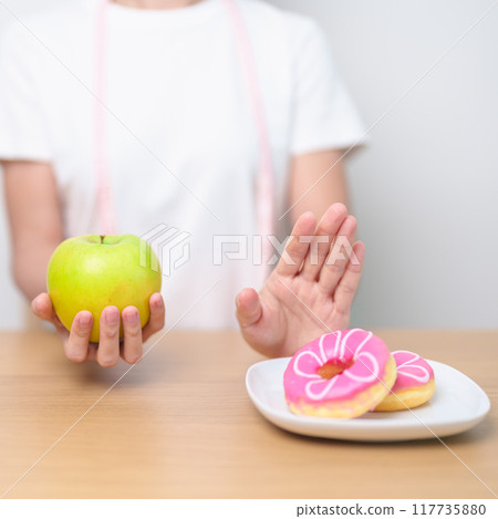 woman hand hold green Apple and reject donut, female fitness choose between fruit is Healthy and sweet is Unhealthy junk food. Dieting control, Weight loss, Obesity, eating lifestyle and nutrition 117735880