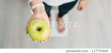 woman on weight scale and hand hold green Apple, choose fruit is Healthy food. Dieting control, Weight loss, Obesity, eating lifestyle and nutrition concepts 117735884