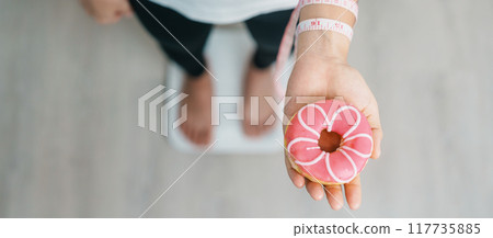 woman on weight scale and hand hold pink Donut, choose stop eating sweet is Unhealthy ealthy food. Dieting control, Weight loss, Obesity, eating lifestyle and nutrition concepts woman on weight scale and hand hold pink Donut, choose stop eating sweet is Unhealthy ealthy food. Dieting control, Weight loss, Obesity, eating lifestyle and nutrition concepts 117735885