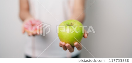 woman hand hold green Apple and donut, female fitness choose between fruit is Healthy and sweet is Unhealthy junk food. Dieting control, Weight loss, Obesity, eating lifestyle and nutrition concept 117735888