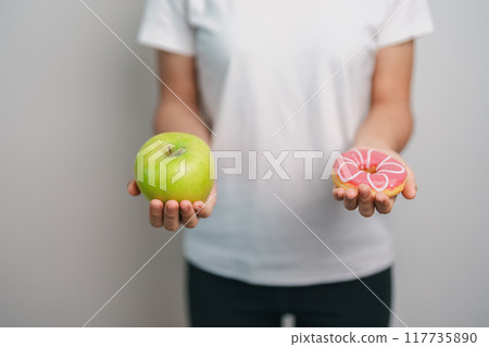 woman hand hold green Apple and donut, female fitness choose between fruit is Healthy and sweet is Unhealthy junk food. Dieting control, Weight loss, Obesity, eating lifestyle and nutrition concept 117735890