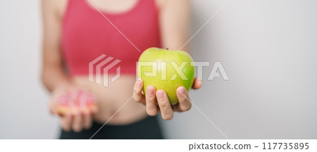 woman hand hold green Apple and donut, female fitness choose between fruit is Healthy and sweet is Unhealthy junk food. Dieting control, Weight loss, Obesity, eating lifestyle and nutrition concept 117735895