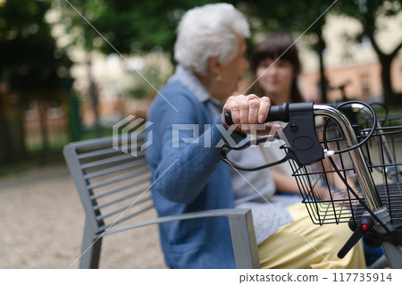 Grandma with walker spending time with granddaughter in city park, sitting on bench at children playground, talking. After school. Grandma with walker spending time with granddaughter in city park, sitting on bench at children playground, talking. After school. 117735914