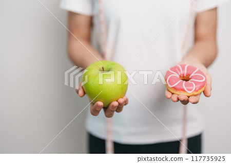 woman hand hold green Apple and donut, female fitness choose between fruit is Healthy and sweet is Unhealthy junk food. Dieting control, Weight loss, Obesity, eating lifestyle and nutrition concept 117735925