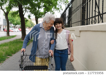 Grandma picking up girl after first day of school. Granddaughter spending time with senior grandmother outdoors in city, walking. Grandma picking up girl after first day of school. Granddaughter spending time with senior grandmother outdoors in city, walking. 117735956