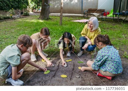 Students playing tic-tac-toe with stones and leaves, grid in soil. Outdoor sustainable education class. 117736031